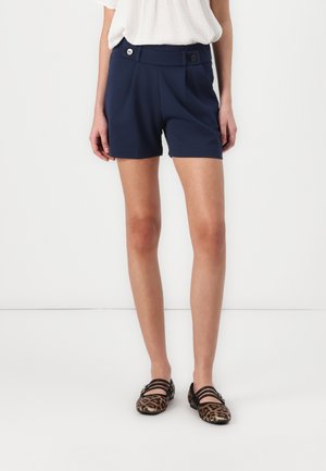 Femme portant un short taille haute bleu marine avec des détails de boutons, un chemisier blanc et des chaussures plates à imprimé léopard avec des brides noires.