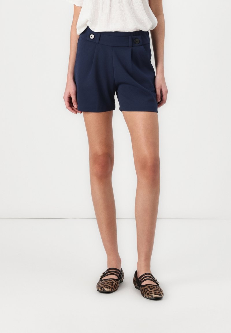Femme portant un short taille haute bleu marine avec des détails de boutons, un chemisier blanc et des chaussures plates à imprimé léopard avec des brides noires.