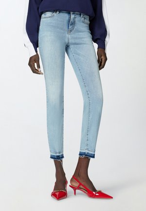 Jeans skinny blu chiaro con orlo sfilacciato, abbinati a décolleté rosse lucide con punta a freccia e dettagli in catena.