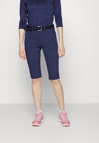 Mørkeblå knelange shorts, kombinert med en mørk mønstret topp og lyse rosa sporty sko med hvite såler. En svart vevd belte fremhever antrekket.