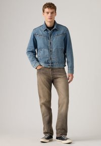 Νέος άνδρας στέκεται με τα χέρια στις τσέπες, φορώντας μπλε τζιν μπουφάν, ξεθωριασμένο καφέ τζιν και μαύρα παπούτσια τύπου low-top, σε απλό φόντο.