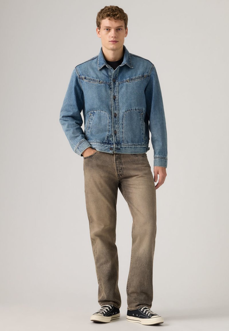 Joven de pie con las manos en los bolsillos, vistiendo una chaqueta de mezclilla azul, jeans marrones descoloridos y zapatillas negras bajas sobre un fondo liso.