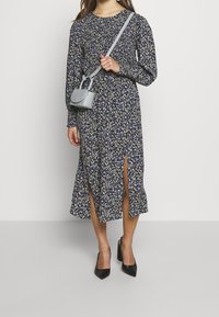 Laivastonsininen pitkähihainen mekkko, jossa on kukkakuosi ja edessä halkiot, yhdistettynä pieneen harmaaseen käsilaukkuun ja mustiin korkokenkiin.