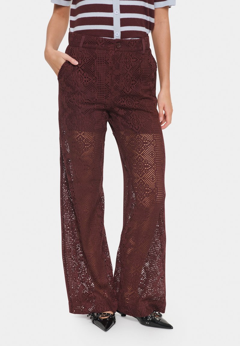Pantaloni wide-leg in pizzo bordeaux con un dettagliato motivo a intaglio, caratterizzati da una vita alta e tasche laterali, abbinati a scarpe nere a punta.