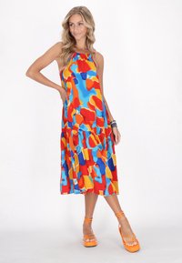 Farbenfrohes Maxikleid mit roten, blauen und gelben abstrakten Mustern, Neckholder-Ausschnitt und gestuften Design. Getragen mit orangen Riemchen-Schuhen.