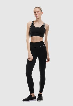 Czarny sportowy biustonosz z białymi akcentami, czarne legginsy z wysokim stanem oraz czarne buty sportowe. Dopasowany krój, gładka tekstura.