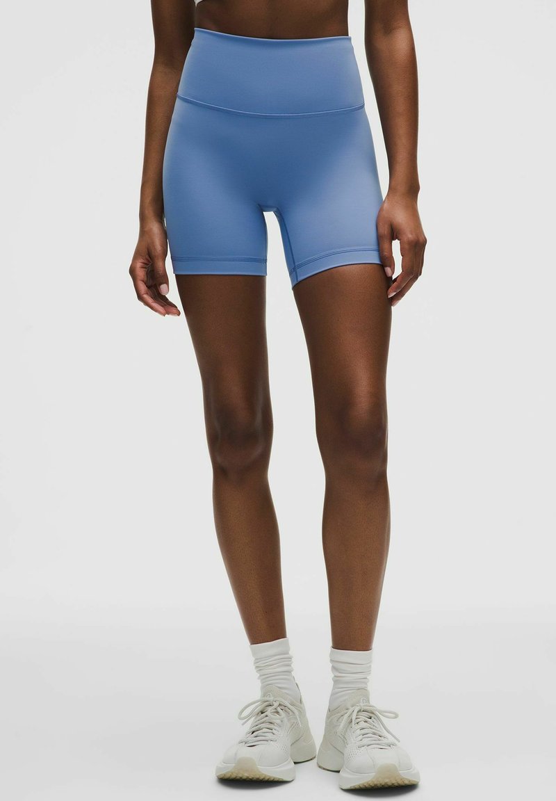 Kvinde iført lyseblå, højtaljede atletiske shorts, hvide sokker og hvide sneakers, stående mod en ensfarvet baggrund.