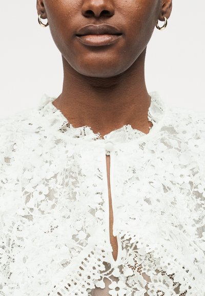 Gros plan d'une personne portant un chemisier en dentelle blanche à motifs floraux, un col montant à volants et de petites boucles d'oreilles créoles.