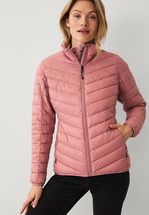 Vrouw draagt een lichtroze gewatteerde jas met een ritssluiting aan de voorkant, staand met één hand in haar zak tegen een effen lichte achtergrond.