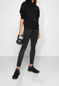 Persona che indossa un hoodie nero, leggings e sneakers, mentre tiene un kettlebell nero con striscia blu su uno sfondo bianco uniforme.