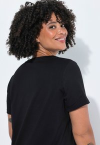 Czarny bawełniany t-shirt z krótkimi rękawami, okrągłym dekoltem i luźnym krojem. Widoczne szczegóły naturalnych loków i faktury skóry.