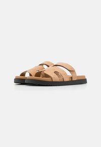 Sandalen aus beigem Wildleder verfügen über kreuzverzurrte Riemen, flache schwarze Gummisohlen und ein minimalistisches Design mit einer offenen Zehenstruktur.