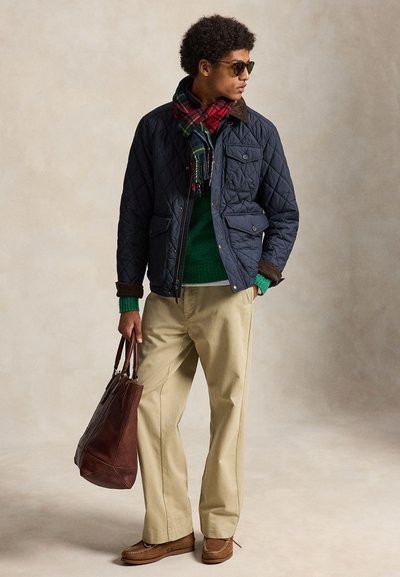 Homme portant une veste matelassée bleu marine, un pull vert, un pantalon beige, une écharpe à carreaux rouge, des lunettes de soleil, des chaussures marron, tenant un grand sac en cuir marron.