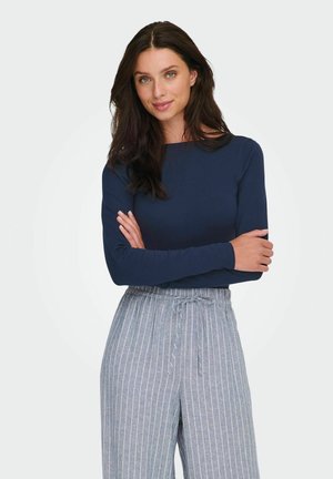 Kobieta o długich ciemnych włosach, ubrana w granatową bluzkę z długim rękawem i szare spodnie w prążki z sznurkiem, stojąca z założonymi na siebie ramionami.