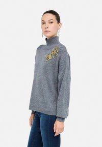 Vrouw in een grijze coltrui met gouden kristalversiering op de schouder, gecombineerd met donkerblauwe jeans, staand tegen een witte achtergrond.