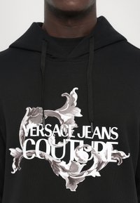 Hoodie preto feito de uma mistura de algodão, apresentando uma grande ilustração floral em branco e cinza e o texto "VERSACE JEANS COUTURE" na parte da frente.
