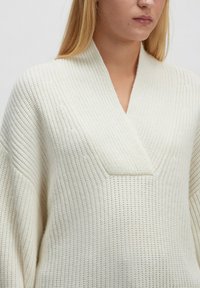 Cremefarbener gestrickter Pullover mit V-Ausschnitt, weiten Ärmeln und strukturiertem Stoff, der ein zentrales Paneel-Detail aufweist.