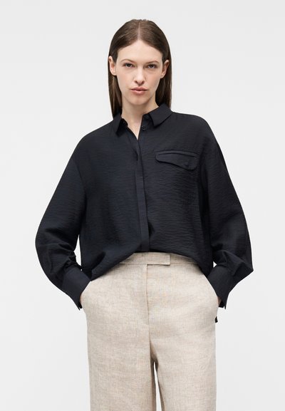 Γυναίκα που φοράει μαύρο πουκάμισο με κουμπιά και μακριά μανίκια με υφή, στερεωμένο μέσα σε μπεζ παντελόνι ψηλής μέσης με φαρδιά πόδια, με τα χέρια στις τσέπες.