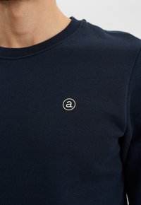 Jersey de cuello redondo azul marino con una textura suave, que presenta un logotipo circular con la letra "a" en el área del pecho izquierdo.