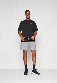 Camiseta negra de manga corta con un pequeño logo, combinada con pantalones cortos deportivos grises. El modelo sostiene una botella de agua negra, vistiendo zapatillas y calcetines negros.