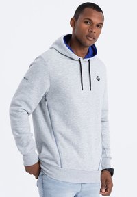 Sudadera gris con un forro interior azul, que cuenta con un bolsillo lateral con cremallera, cordones ajustables negros y un parche con logo en el pecho.