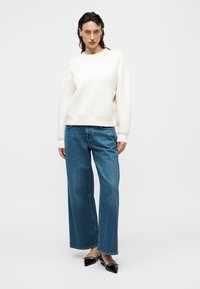 Femme debout, portant un sweat-shirt blanc à manches longues, un jean bleu à jambes larges et des chaussures plates noires à bout pointu, sur un fond clair uni.