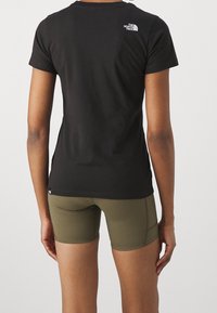 Persona che indossa una maglietta nera The North Face e pantaloni corti aderenti verdi oliva, mostrata di spalle su uno sfondo uniforme.