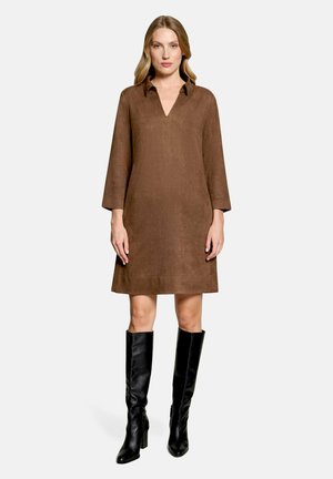 Frau trägt ein braunes Langarmkleid mit Kragen und V-Ausschnitt sowie schwarze kniehohe Stiefel mit Absatz und steht vor einem schlichten Hintergrund.