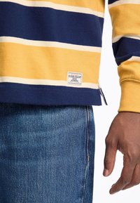 Yellow and navy striped sweatshirt with a woven "Guess Jeans" label, paired with blue denim jeans. Features side slits and ribbed cuffs.