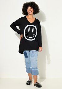 Tunic nera in maglia con grafica di una faccina sorridente bianca, abbinata a jeans cropped in denim azzurro chiaro con strappi, diverse toppe e orli sfilacciati.