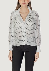 Blusa bianca plissettata con pois neri, chiusura con bottoni e maniche lunghe con polsini a contrasto. Il tessuto trasparente aggiunge texture.