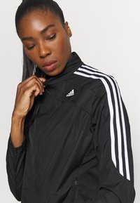 Giacca a vento nera Adidas con colletto alto, accenti bianchi a tre strisce sulle spalle e una tasca laterale con zip. Tessuto morbido e testurizzato.