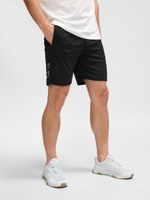 Mann trägt schwarze Shorts mit weißem geometrischem Logo an der Seite und weiße Sneakers, steht mit Händen in den Taschen vor grauem Hintergrund.