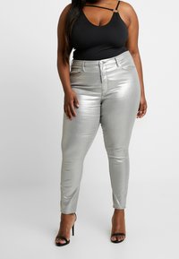 Jeans ajustados metálicos plateados con una cintura alta y bolsillos delanteros. Top negro sin mangas con un diseño de tirantes. Sandalias negras de tacón.