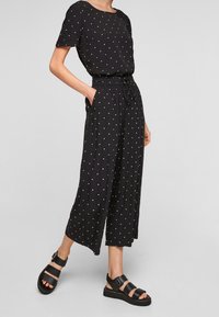 Combinaison noire à pois avec manches courtes, encolure ronde, taille élastique et jambes larges, associée à des sandales noires à boucles.