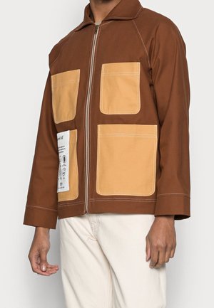 Hnědá bunda na zip se čtyřmi velkými světle hnědými kapsami, z nichž jedna má bílou etiketu s černým textem a ikonami, nošená s světlými kalhotami.