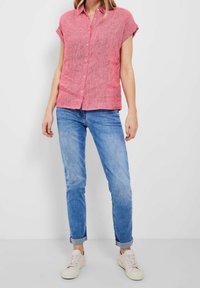 Mujer vestida con una camisa de botón rosa de manga corta, jeans azules remangados y zapatillas deportivas blancas, de pie contra un fondo blanco liso.