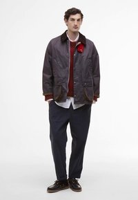 Άνδρας που φοράει σκούρο γκρι μπουφάν πάνω από κόκκινο πουλόβερ και λευκό πουκάμισο, ναυτικές παντελόνες και μαύρα κορδόνια παπούτσια, στέκεται μπροστά σε λευκό φόντο.