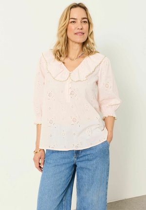 Frau mit blonden Haaren, die eine hellrosa Bluse mit großem gewelltem Kragen und blaue Jeans trägt, steht vor einem schlichten hellen Hintergrund.