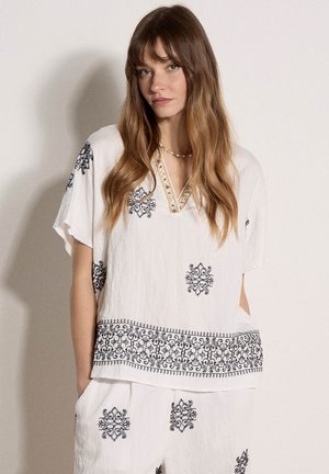 Mujer vestida con blusa blanca holgada y pantalones con patrones bordados en negro, de pie contra un fondo claro y sencillo.