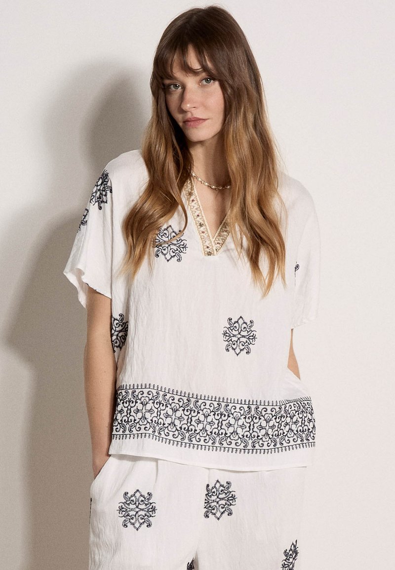 Femme portant un haut blanc ample et un pantalon avec des motifs brodés noirs, debout devant un fond clair uni.