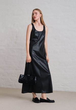 Une femme présente une longue robe noire effet cuir, porte un sac à main noir et des mocassins noirs devant un mur de briques clair.