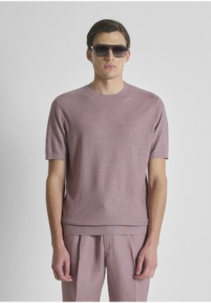Uomo che indossa un maglione viola chiaro a maniche corte, pantaloni abbinati e occhiali da sole grandi e scuri, in piedi davanti a uno sfondo chiaro uniforme.