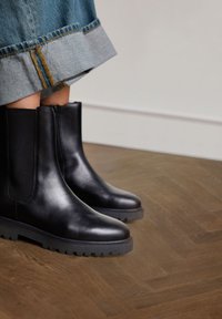 Zwarte leren enkellaarzen met elastische zijpanelen en getextureerde rubberen zolen, geplaatst op een houten vloer, gedeeltelijk bedekt door denim.