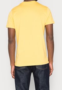 Camiseta de algodón amarilla con mangas cortas, cuello redondo y un ajuste relajado. Llevada con jeans de mezclilla oscura, mostrando mínimos detalles en la parte trasera.