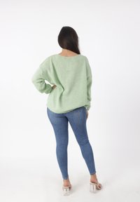 Hellgrüner, strukturierter Pullover mit lockerer Passform, runder Ausschnitt und überschnittenen Schultern; kombiniert mit enganliegenden blauen Jeans und transparenten Sandalen mit Absatz.