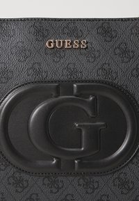 Musta tekstuurihoonega kott, millel on tõstetud must topelt G logo läikiv märk ja kuldne Guess brändinimi koti ülemises osas.