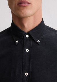 Schwarzes Cordhemd mit einem Button-Down-Kragen, ausgestattet mit weißen Knöpfen und einer strukturierten Oberfläche. Nahaufnahme, die auf Details fokussiert.