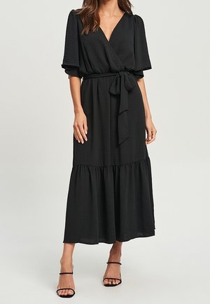 Mujer que lleva un vestido envolvente negro con cintura atada, mangas abullonadas hasta el codo y una falda de volantes, combinado con tacones negros con tiras.