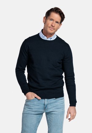 Man met kort bruin haar draagt een lichtblauw overhemd onder een zwarte trui en lichtblauwe spijkerbroek, staand met één hand in de zak.